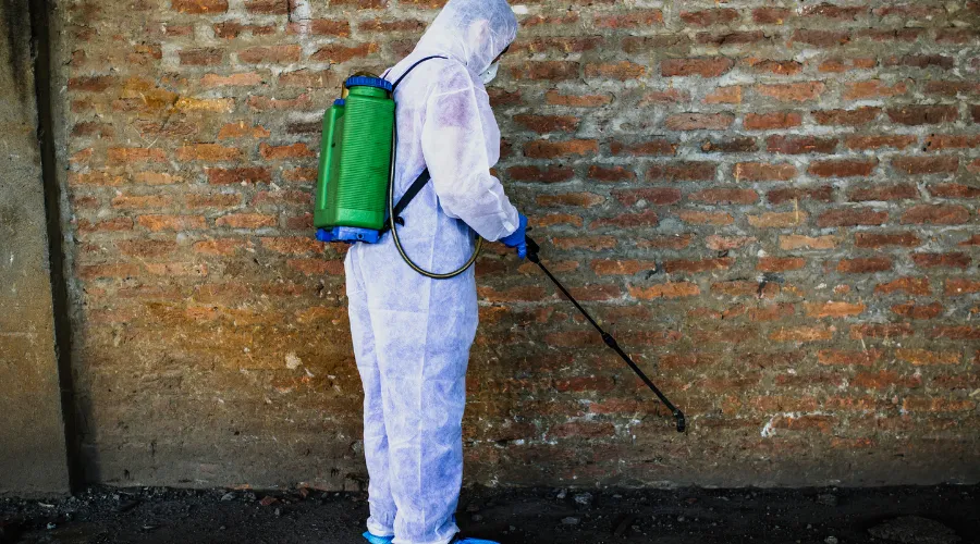 Person in a full hazmat suit spraying a brick wall with a green backpack sprayer on the exterior surface and ground nearby.