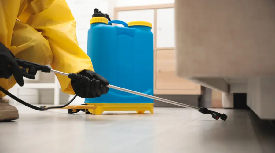 Person in a yellow hazmat suit spraying disinfectant with a blue backpack sprayer on the floor.